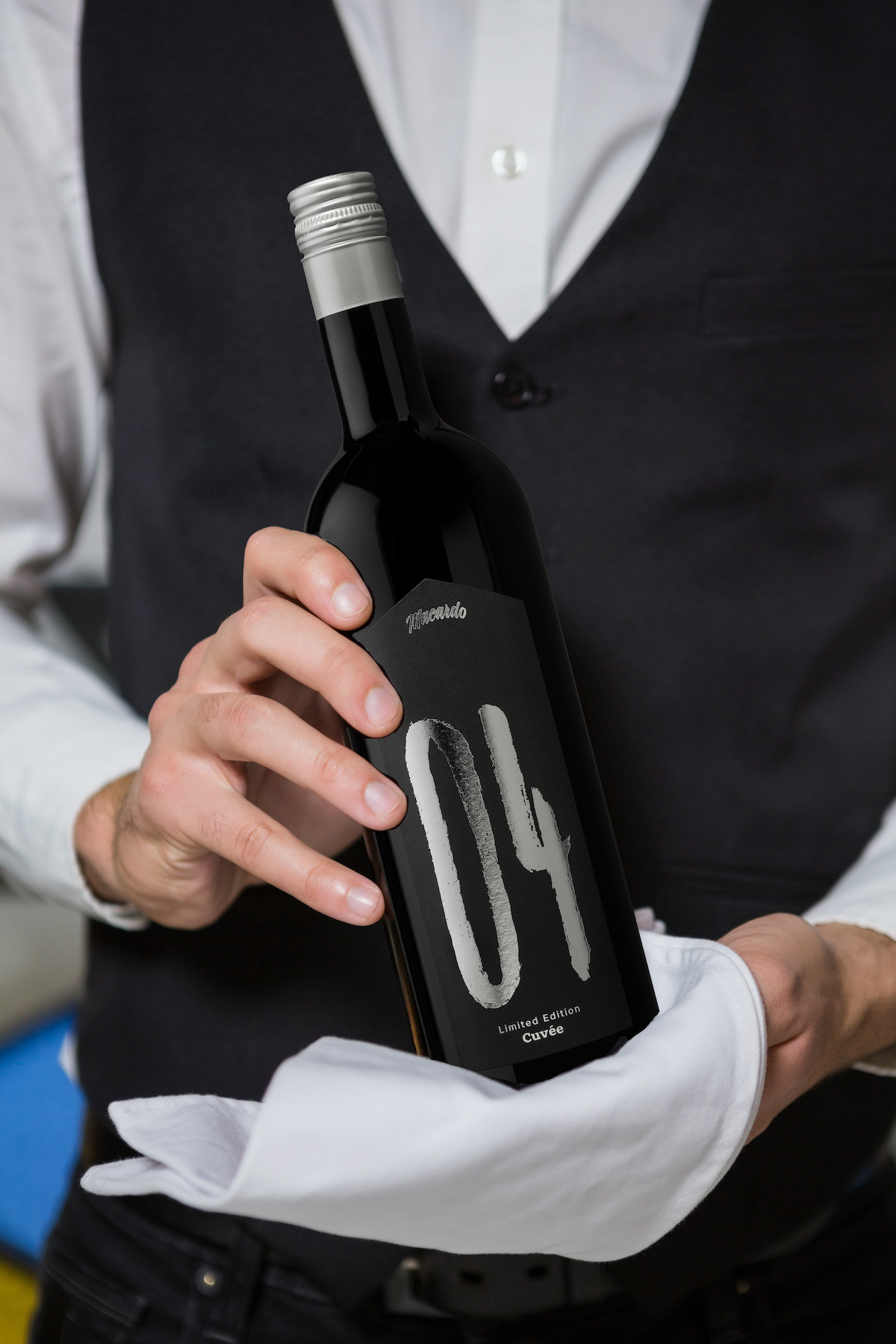 Ein Mann im Anzug hält eine dunkle Weinflasche mit silbernem Schraubverschluss in einem weißen Tuch. Das Etikett der Flasche zeigt die silberne Zahl "04" und den Schriftzug "Limited Edition Cuvée".